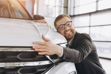 Positive young bearded caucasian man in glasses hugging his new car in dealership. Happy man finally gets long-awaited car, wearing formal suit