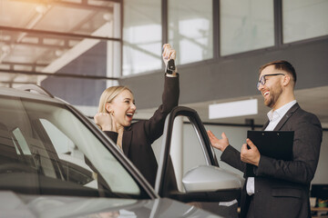 Modern happy woman raising hand up making fist near her new car in car showroom. Bearded seller consultant in a suit