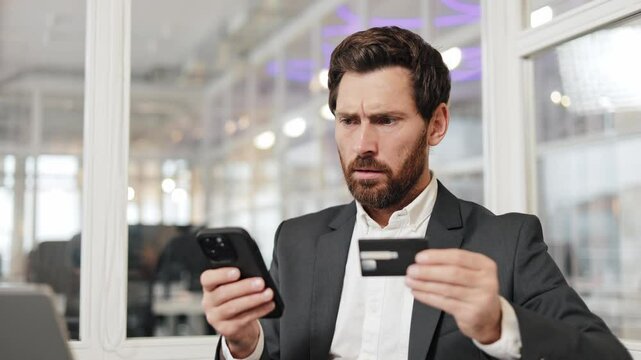 A businessman in a suit is experiencing problems with online payment. He is holding his phone and credit card, looking puzzled and frustrated. His facial expression suggests confusion and worry.