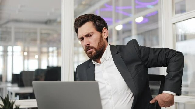 Office worker businessman sits at his desk and works on his laptop. Man develops back pain and massages his lower back. Office work can be physically demanding.