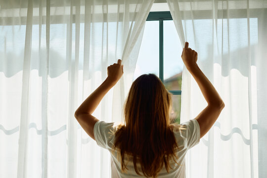 Rear view of woman opening curtains on window in  morning.
