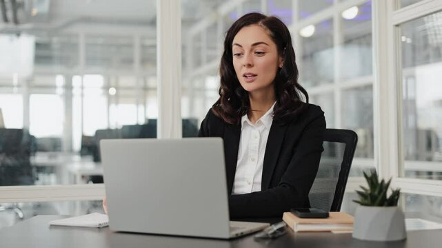 Serious businesswoman in a suit sits at her desk in an office, using a laptop for a video conference call. Corporate leader talks to the camera, taking notes on a notepad.