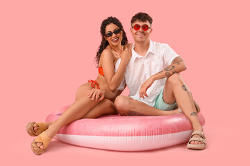 Happy young couple in sunglasses sitting on inflatable mattress against pink background