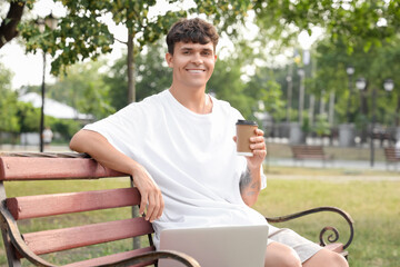 Handsome young man with laptop and cup of delicious latte sitting on bench outdoors