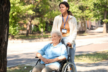 Young African-American female medical worker walking with elderly woman on wheelchair outdoors