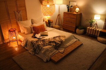 Interior of bedroom with glowing lamps, laptop and books on bed in evening