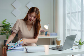 Young Asian woman looking happy with headphones around her neck Work on a laptop computer online business Communicate with video conferencing Take notes in a notebook Ideas at the work desk.