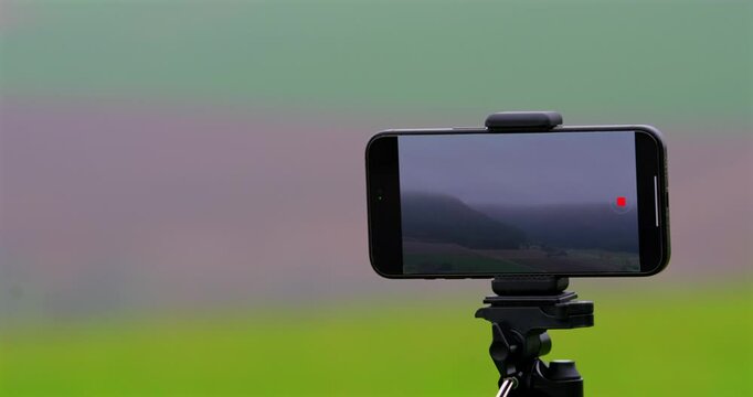 A smartphone affixed to a tripod records a foggy landscape, offering a lens into the mist-enveloped fields and distant forests with quiet dedication.