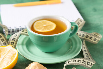 Cup of tea for weight loss, notebook and measuring tape on green table, closeup