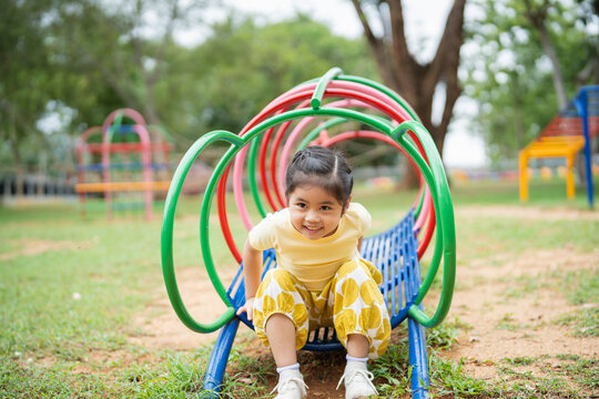 Asian young girl is sitting on a slide in a park. She is smiling and looking up at the camera. The slide is surrounded by other playground equipment, including a swing set and jungle gym