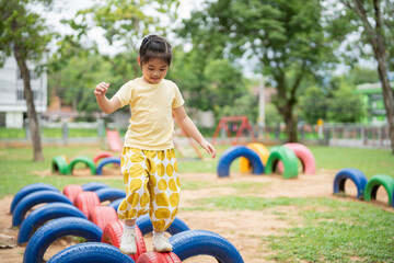 Asian young girl is playing in a park with a tire obstacle course. She is wearing a yellow shirt and yellow pants