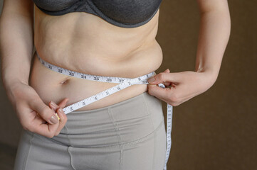 Overweight woman with belly fat carefully measures her waist with tape measure to make sure the...