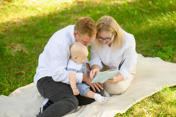 Caring parents read an interesting book with children stories and fairy tales to child, sitting on blanket during picnic in park on sunny day, showing baby pictures. Communication parents and toddler