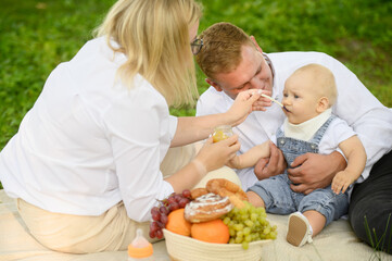 Dad holds baby in his arms, mom feeds happy baby healthy food from jar with spoon, during picnic in park, close up. Baby tries vegetable mix from jar. Healthy eating concept, happy family.