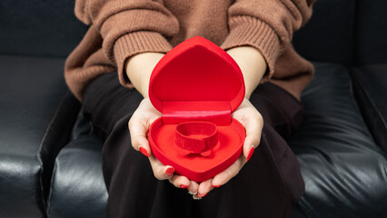 Asian woman sitting on black sofa and holding red heart shaped gift box with gold bow in hand. Gift box open lid. Presents and gift boxes for festival and celebration.