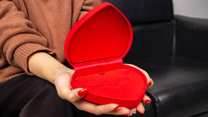 Asian woman sitting on black sofa and holding red heart shaped gift box with gold bow in hand. Gift box open lid, present and gift boxes for festival and celebration.