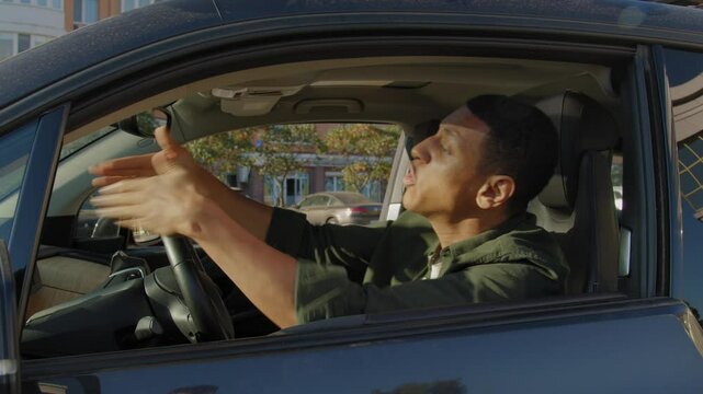 Angry irritated handsome black male driver stuck in traffic jam, beeping car horn, peeking out from car window, gesturing and arguing while driving vehicle in the city on busy road.