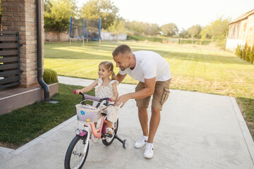 Supportive young father is helping his daughter to learn how to ride a bicycle in backyard.