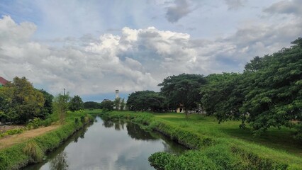 Fototapeta premium A serene riverside scene with lush greenery under a cloudy sky. The calm river reflects the surrounding trees, while a tall tower stands in the distance, adding depth to the peaceful landscape.