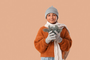 Cute African-American girl in stylish warm clothes with Christmas tree branches on brown background