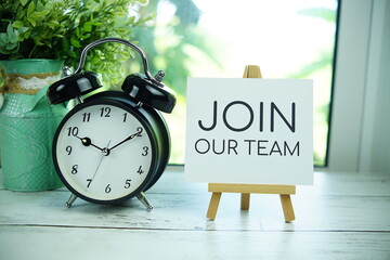 Join Our Team quotes text on paper card with easel and alarm clock in the window background