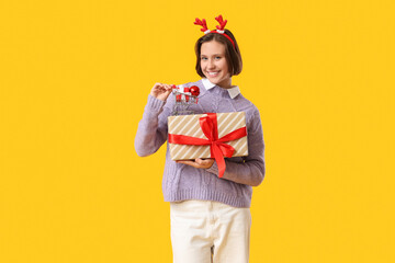 Beautiful young happy woman in reindeer horns with gift boxes and shopping cart on yellow background. Christmas celebration