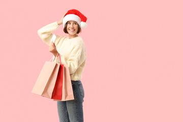 Beautiful young happy woman in Santa hat with shopping bags and credit card on pink background. Christmas celebration