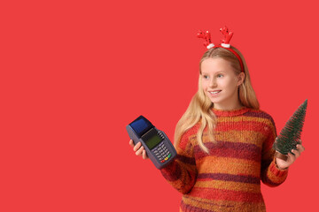 Cute girl in reindeer horns with payment terminal and Christmas tree on red background