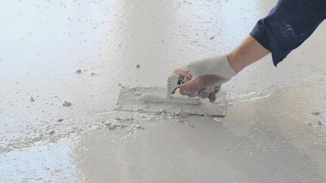 Removal of irregularities and roughness on the concrete floor screed using a metal rectangular spatula. A builder's hand in a glove smoothes concrete with a special tool while it is not yet hardened