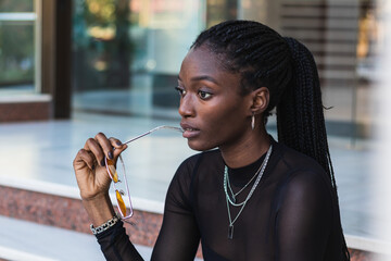 Fashionable young woman with braided hair and sunglasses sits on outdoor steps, deep in thought
