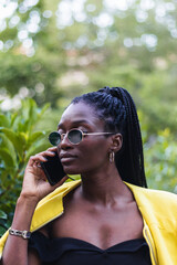 Fototapeta premium Young african american woman in a stylish yellow jacket and sunglasses making a phone call outdoors