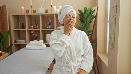 Woman yawning in a spa wearing a white robe and towel on her head in a peaceful, candle-lit room with plants, shelves, and wellness decor in the background