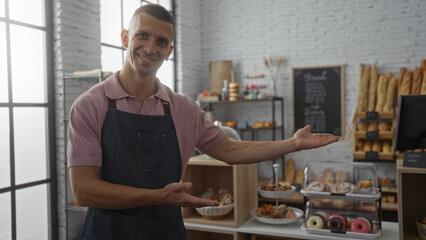 Young man smiling and presenting various pastries displayed in a cozy bakery shop interior with shelves filled with fresh bread and baked goods in the background