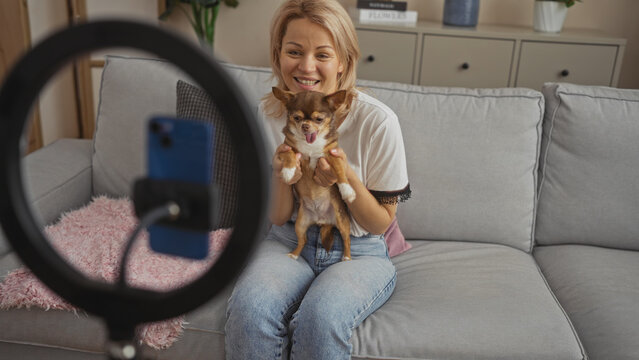 Young blonde woman with a chihuahua dog sitting in a living room, smiling at her phone while recording a video.