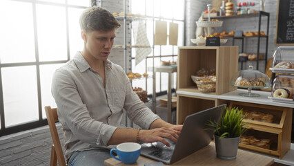 Young man working on laptop in a cozy bakery interior with shelves of pastries and a coffee mug on the table next to him.