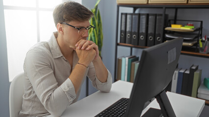 Young man working in an office with a computer and thinking deeply while surrounded by shelves of binders and plants