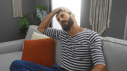 Handsome young hispanic man with a beard sitting indoors on a couch in a living room wearing a striped shirt