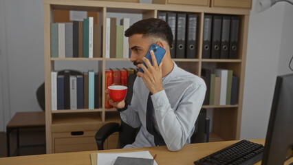 Handsome young hispanic man with a beard holding a coffee cup and talking on the phone in an office interior.