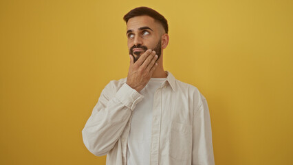 Handsome young hispanic man with a beard standing thoughtfully against an isolated yellow background wall