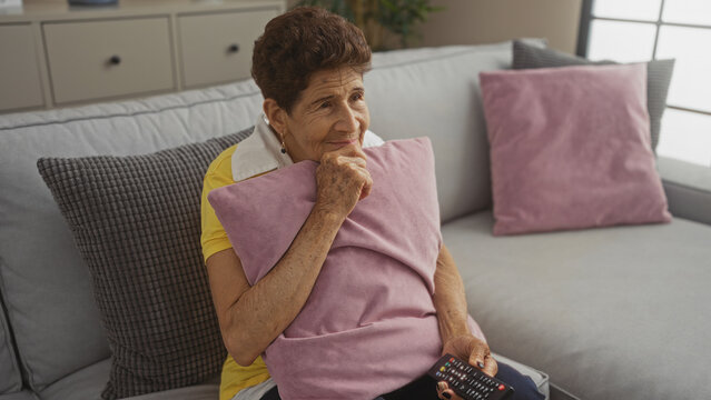 A senior hispanic woman with short hair, in a yellow shirt, holding a remote control and hugging a pink cushion in a cozy living room at home. - Powered by Adobe