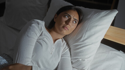 Young hispanic brunette woman indoors in a bedroom lying on a bed with a thoughtful expression on...