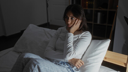 Fototapeta premium Hispanic brunette woman sitting thoughtfully in bed within a dimly lit bedroom of her home, wearing comfortable pajamas and resting against a white pillow.