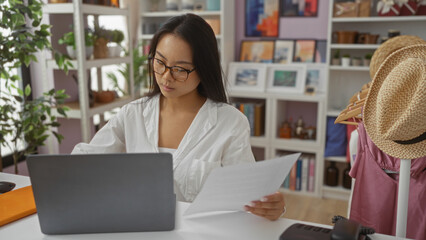 Fototapeta premium Young chinese woman working on a laptop while holding documents in a decorated indoor home decor shop with shelves of plants and various artworks in the background