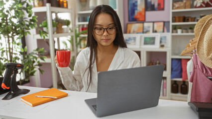 Young chinese woman in an indoor decor shop working on a laptop while holding a red mug surrounded by various home decorations