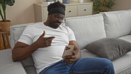 Handsome, young african american man with a beard sitting in a comfortable living room, holding a smartphone, and interacting with it indoors at home.