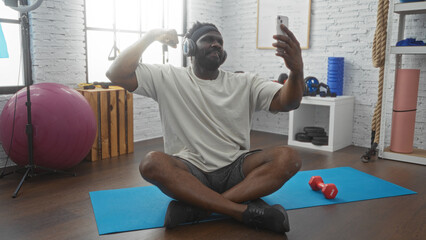 Young african american man with beard exercising indoors in a gym while taking a selfie, sitting on...