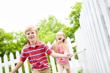 Happy children, siblings or playing with stick in garden for summer together in nature or backyard. Young, brother and sister running with smile for fun childhood, bonding or holiday weekend by fence