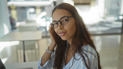 Young woman with glasses sitting in an office interior workspace, looking confidently at the camera, suggesting a professional and engaged atmosphere