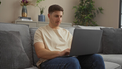 Handsome young hispanic man working on a laptop indoors in a cozy living room of his apartment.