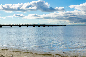 Obraz premium Scenic view of a long wooden pier stretching into calm waters under a cloudy sky near a coastal area during the day